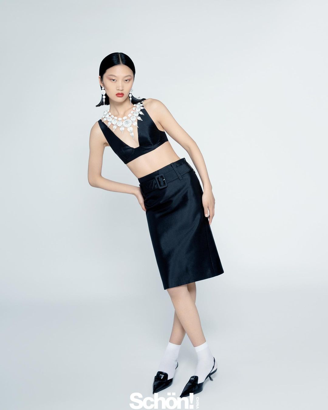 A stylish Asian woman poses indoors wearing a black two-piece outfit, white socks, and black heels against a neutral background.