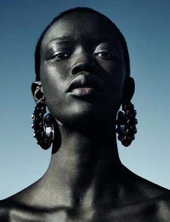 Close-up portrait of a Black woman with short hair wearing large dark studded hoop earrings against a clear blue sky background.