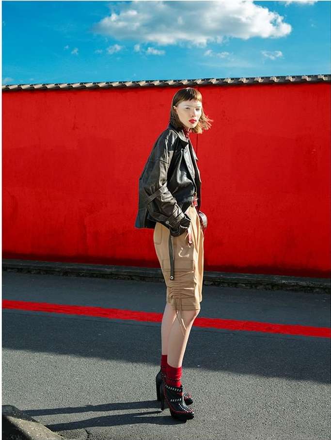 Woman in black jacket and khaki shorts posing in front of a bright red wall against a blue sky on a city street.