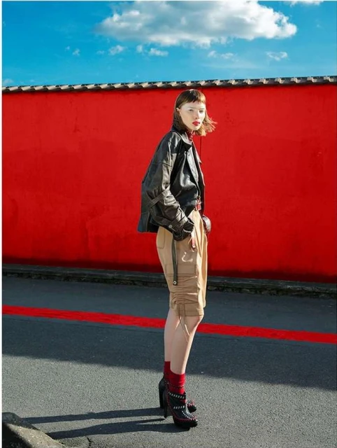 Woman in black jacket and khaki shorts posing in front of a bright red wall against a blue sky on a city street. 