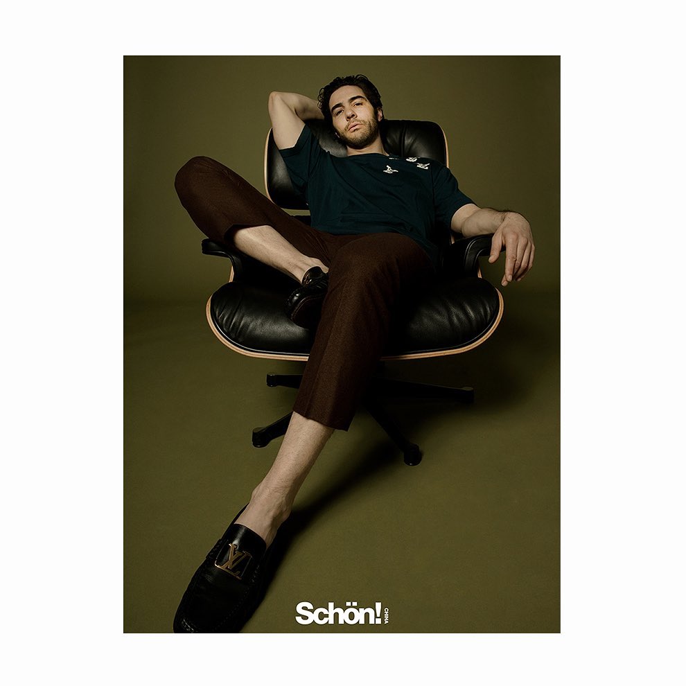 A man relaxes in a black leather chair, wearing a dark green tee shirt and brown pants, captured in a stylish studio fashion portrait.