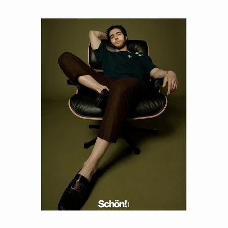 A man relaxes in a black leather chair, wearing a dark green tee shirt and brown pants, captured in a stylish studio fashion portrait.