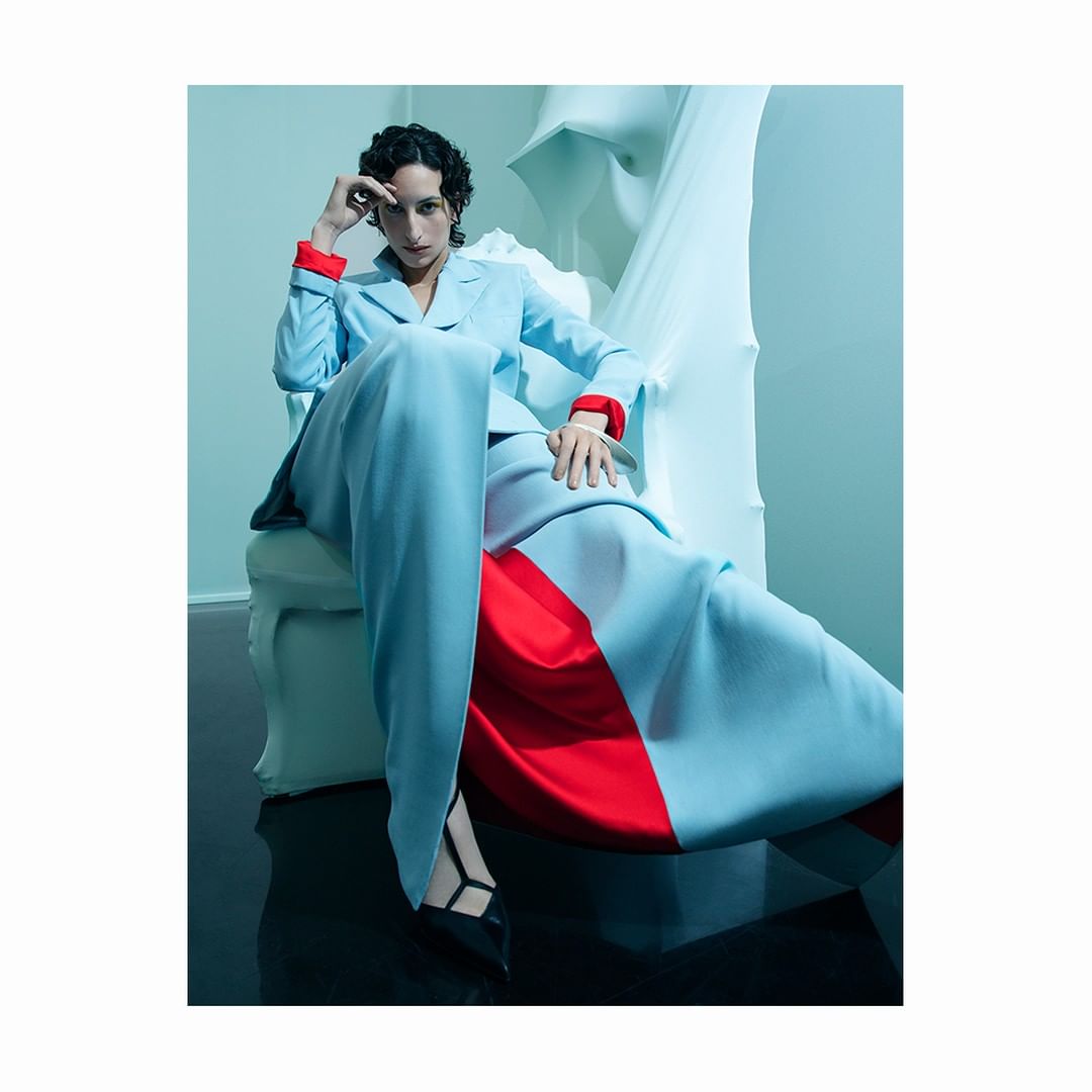 A stylish woman in a blue and red pant suit poses in a photo studio, seated in a white chair against a blue backdrop.