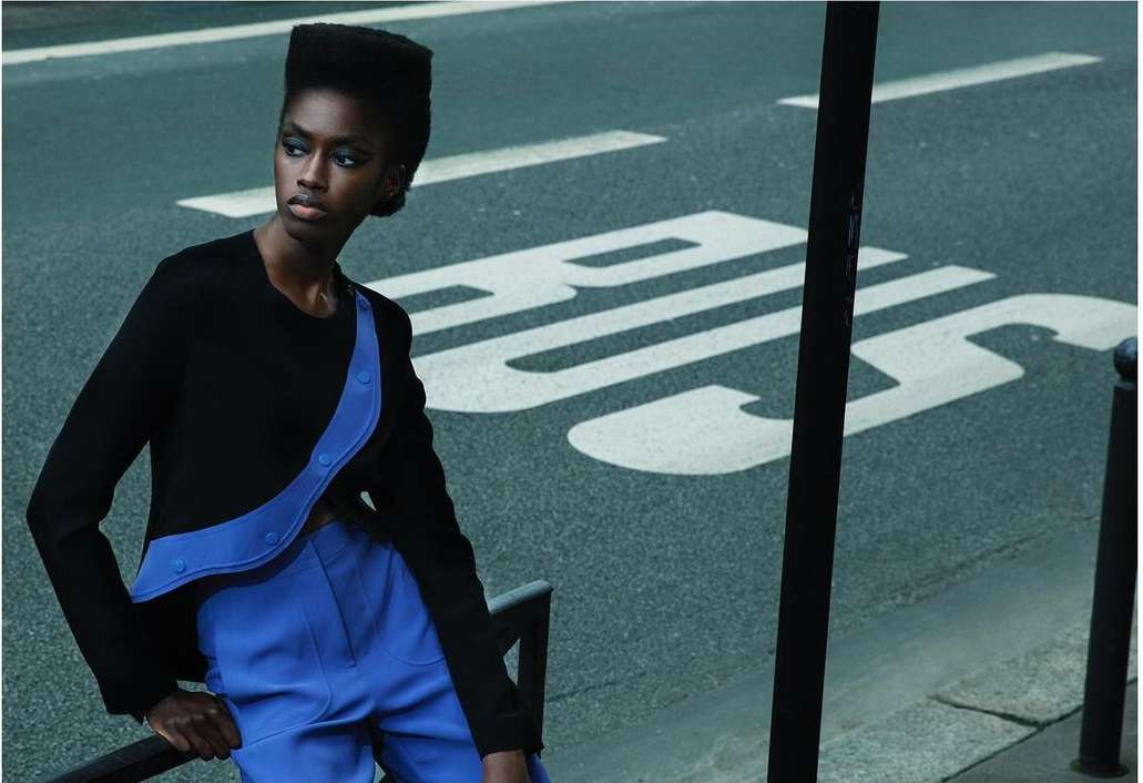 Woman in black and blue fashion stands near bus stop sign on city street, creating a modern, edgy editorial style.
