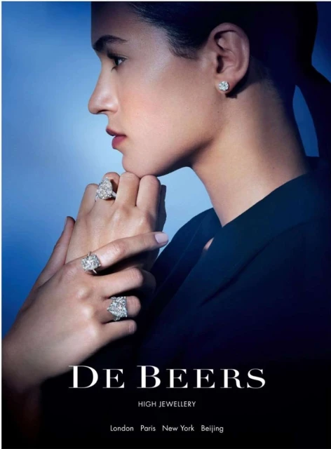 Profile view of a woman with multiple diamond rings and stud earrings against a blue background for De Beers high jewelry ad.