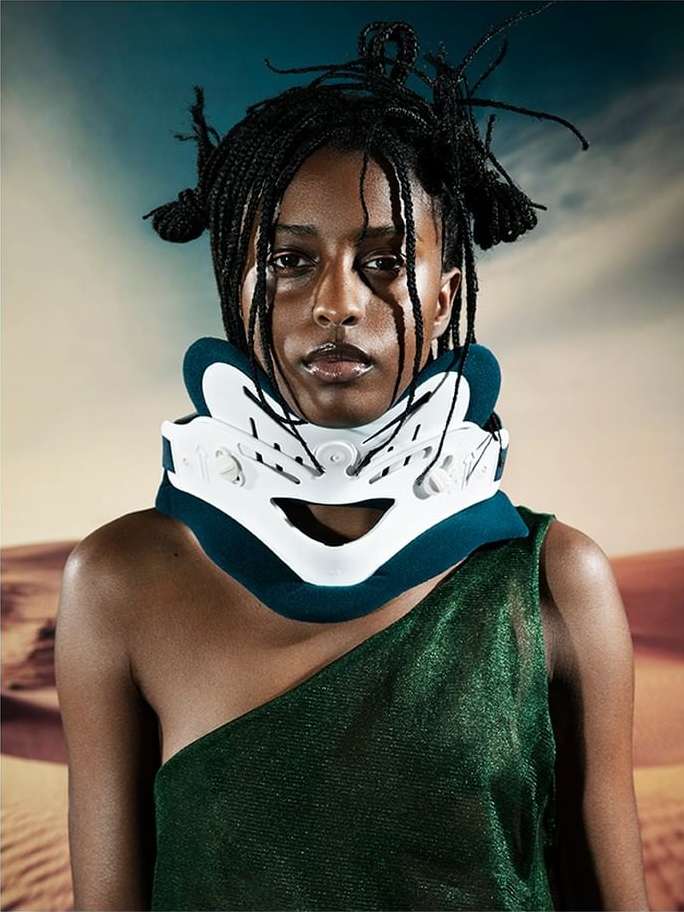 Portrait of a woman wearing a neck brace with an open shoulder dress in front of a desert landscape backdrop, close up studio shot.
