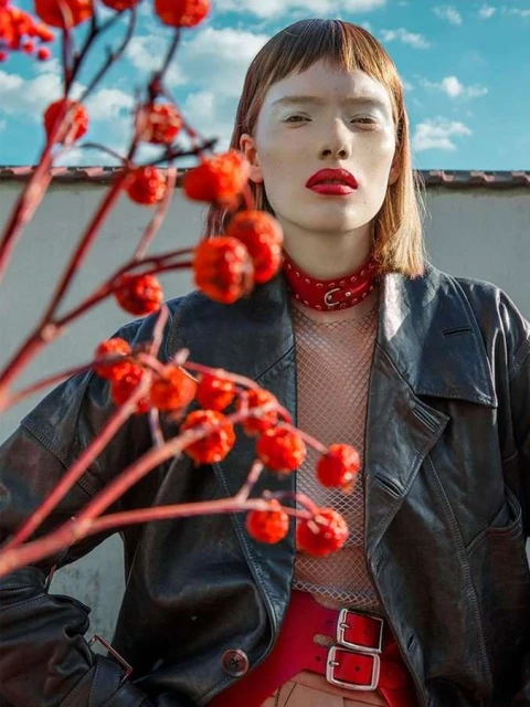 Close-up of a woman with red lipstick and choker in a black leather jacket, accented by out-of-focus red berries against a cloudy sky.