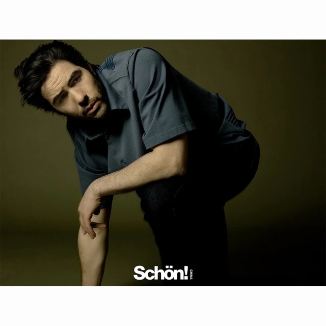 Adult man posing indoors, wearing a short-sleeved button-down shirt and dark trousers, leaning to the side, with a neutral expression.