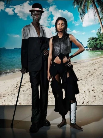 Fashionable couple poses on a beach backdrop. The man wears a two-tone suit and hat, while the woman sports a leather vest and deconstructed pants.