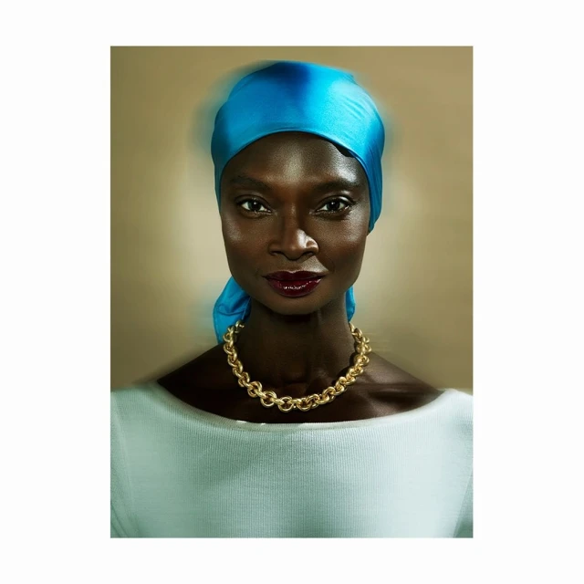 Close-up portrait of a dark-skinned woman with a bright blue headscarf, gold necklace, and white top against a neutral backdrop.