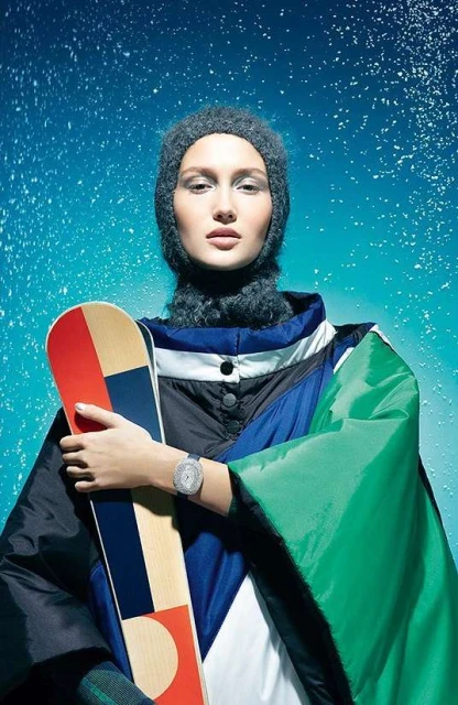 Young woman in winter fashion holding a snowboard against a blue, snowy background. She's wearing a grey beanie and colorful jacket.