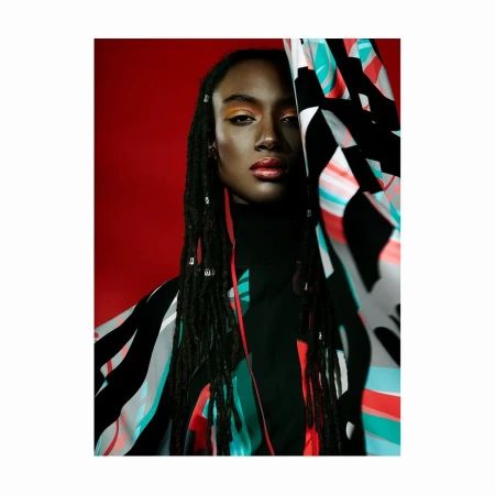 Portrait of a Black woman with dreadlocks and metallic hair cuffs wearing patterned clothing against a solid red background
