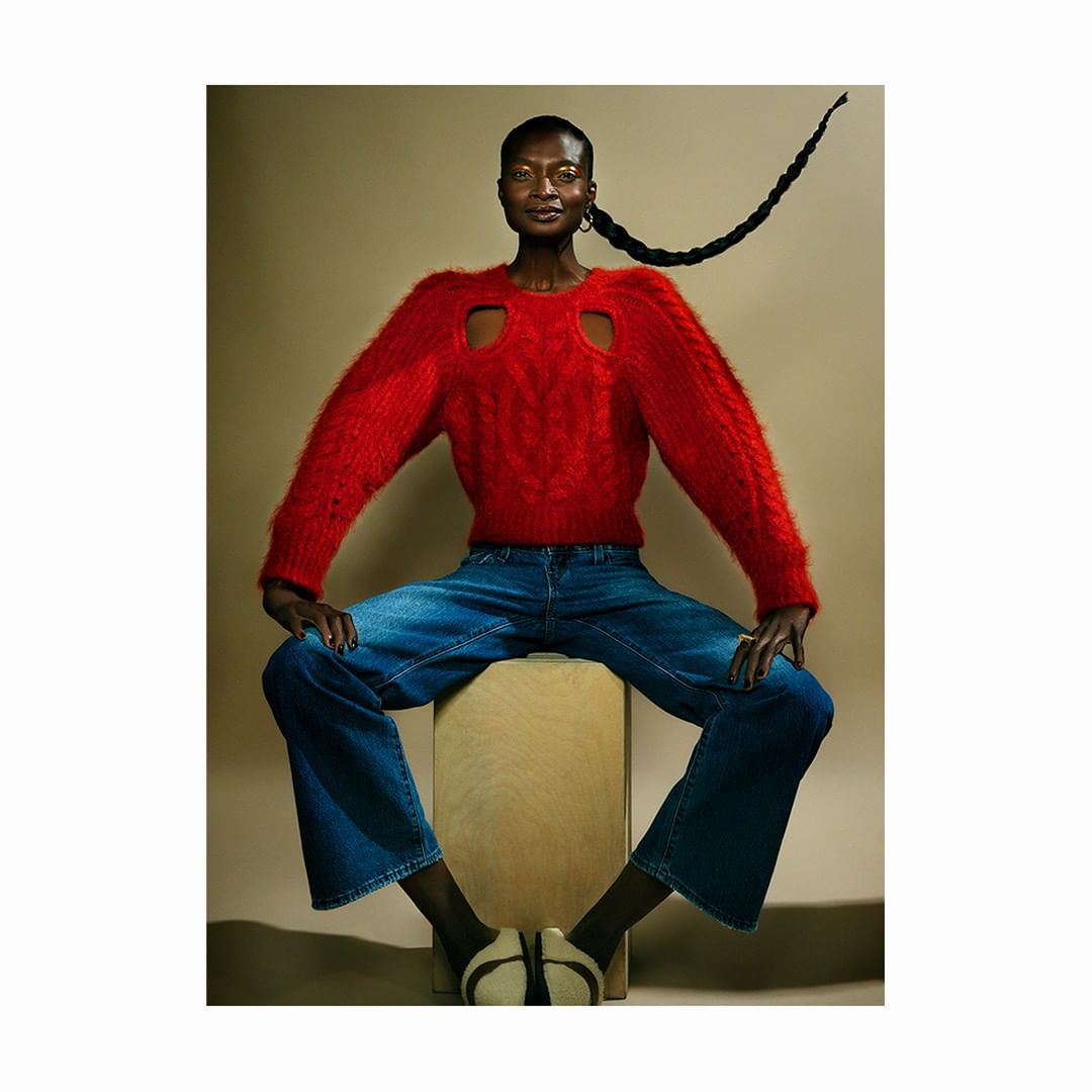 Fashion portrait of Black adult woman in a red sweater and blue jeans, sitting on a wooden box against a neutral background.