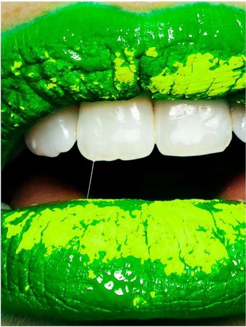 Extreme close-up of a woman's face features vibrant green lipstick, perfectly white teeth, and a single strand of saliva.