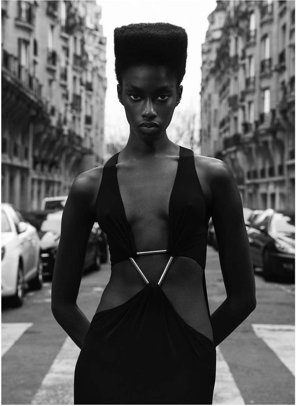 Fashion shot of a Black woman in a black dress with geometric cutouts, posing on a city street with cars and buildings in the background.