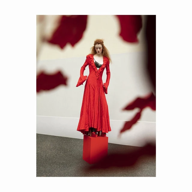 A full shot of a stylish redhead woman in a vibrant red gown with flared sleeves and a ruffled neckline, posing on a red cube.