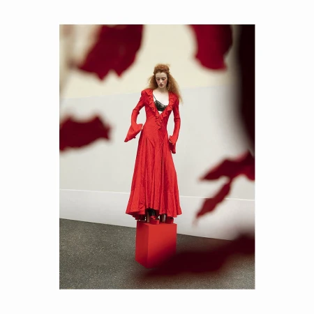 A full shot of a stylish redhead woman in a vibrant red gown with flared sleeves and a ruffled neckline, posing on a red cube.