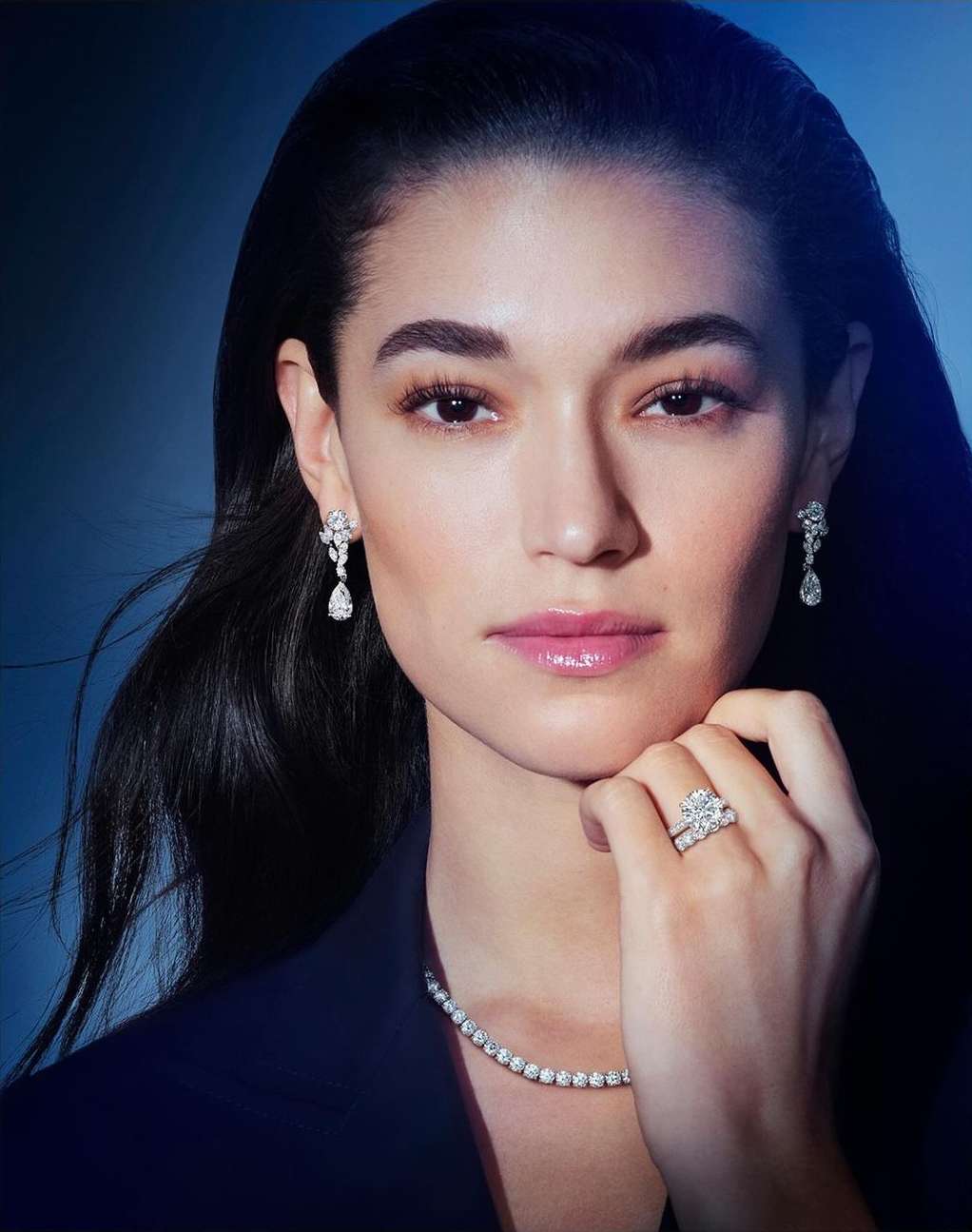 Close-up shot of a stylish adult woman with dark hair wearing diamond earrings, necklace, and ring against a blue-gradient backdrop.