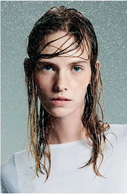 Close-up portrait of a young woman with wet hair, wearing a white shirt, in a studio setting with simulated rain for a fresh beauty look.
