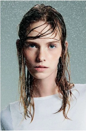 Close-up portrait of a young woman with wet hair, wearing a white shirt, in a studio setting with simulated rain for a fresh beauty look.