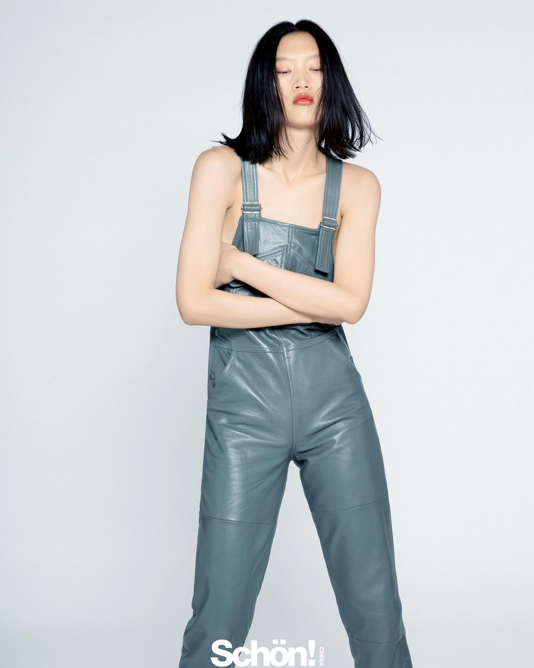 Fashion portrait of an adult woman posing in a studio wearing grey-blue leather overalls and a red lipstick against a white backdrop.