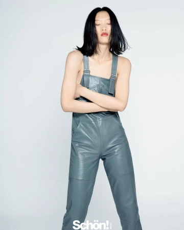 Model with dark shoulder-length hair wearing grey leather overalls against a plain white background