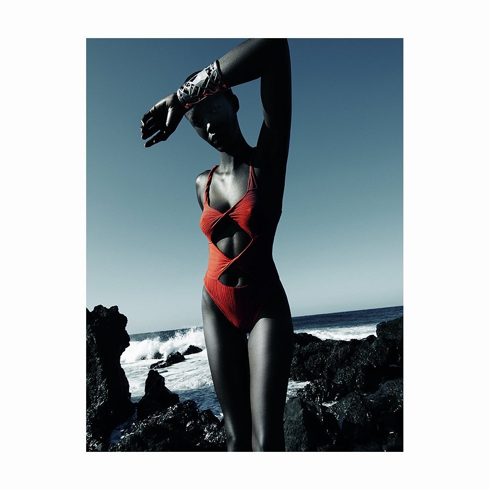 Woman in a red swimsuit poses on a rocky beach against a blue sky and ocean backdrop, showcasing fashion and a coastal setting.