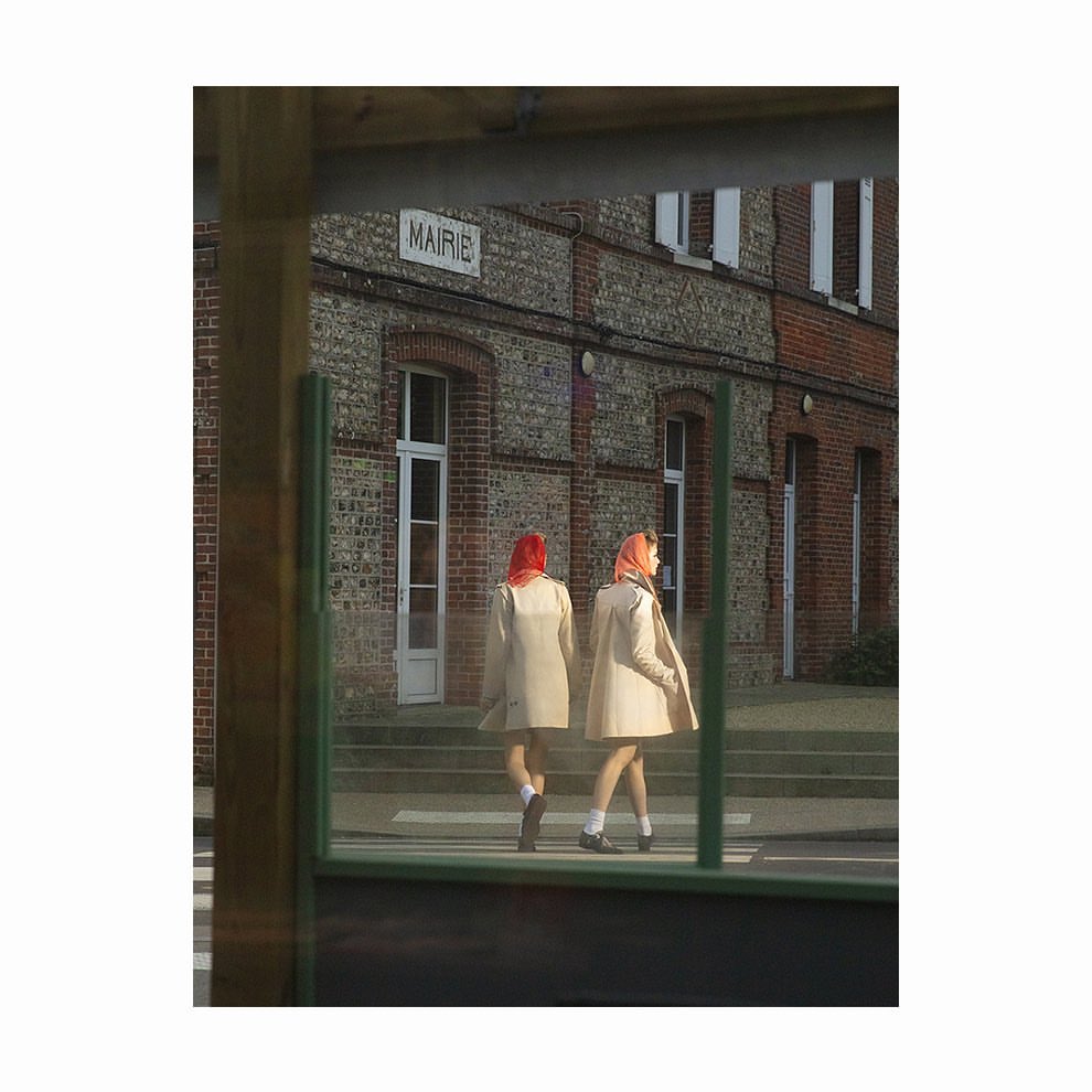 Two women walk past brick building in matching coats and headscarves on an overcast day. Outdoor shot, neutral colours, reflection.