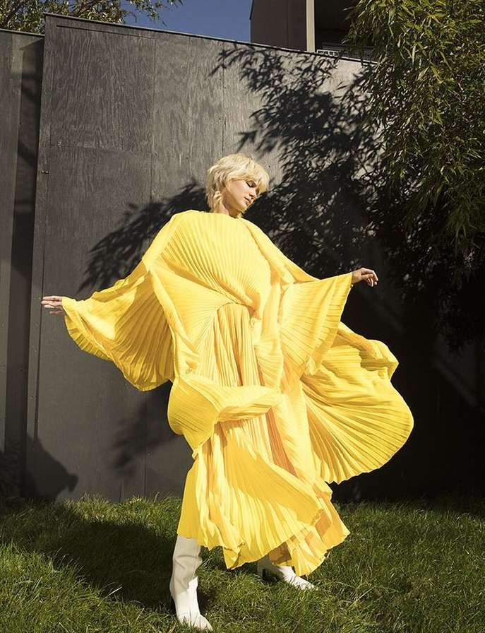 Woman with short blonde hair poses in flowing yellow dress and white boots in front of a dark wall and greenery, creating a striking fashion image.