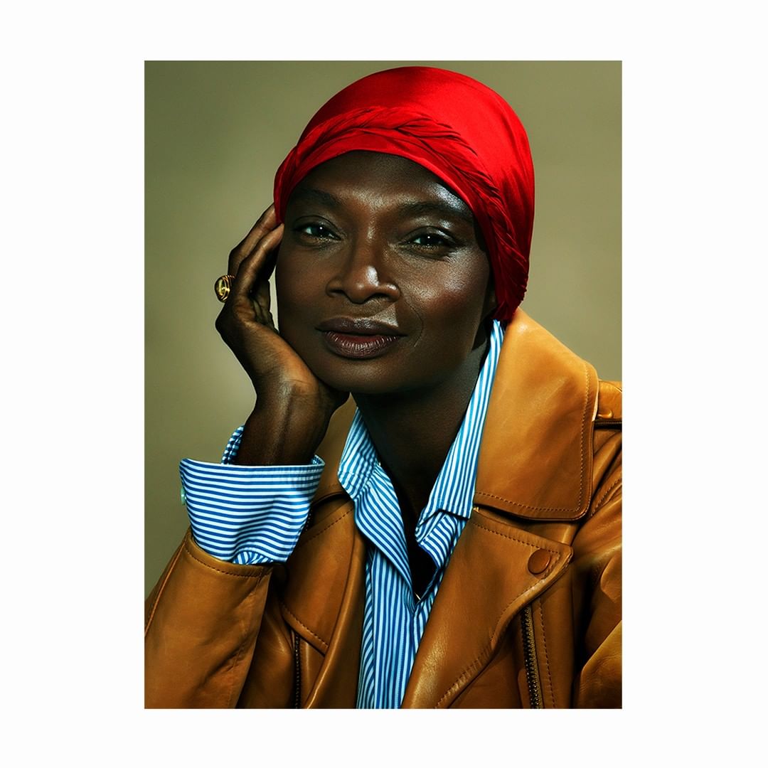 Portrait of a woman with dark skin and red headscarf, wearing a tan leather jacket and striped shirt against a neutral backdrop.