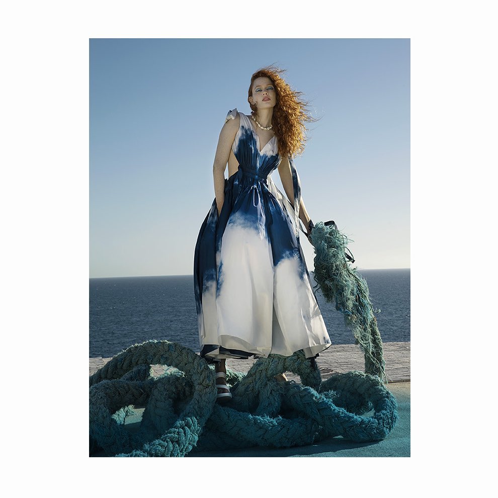 A stylish woman in a blue and white dress stands outdoors by the ocean, holding a thick rope in a fashion editorial shot.