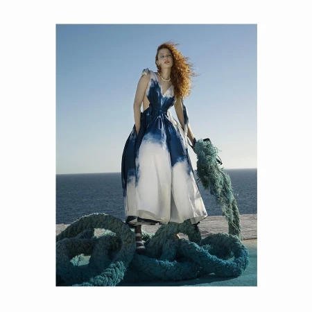 A stylish woman in a blue and white dress stands outdoors by the ocean, holding a thick rope in a fashion editorial shot.