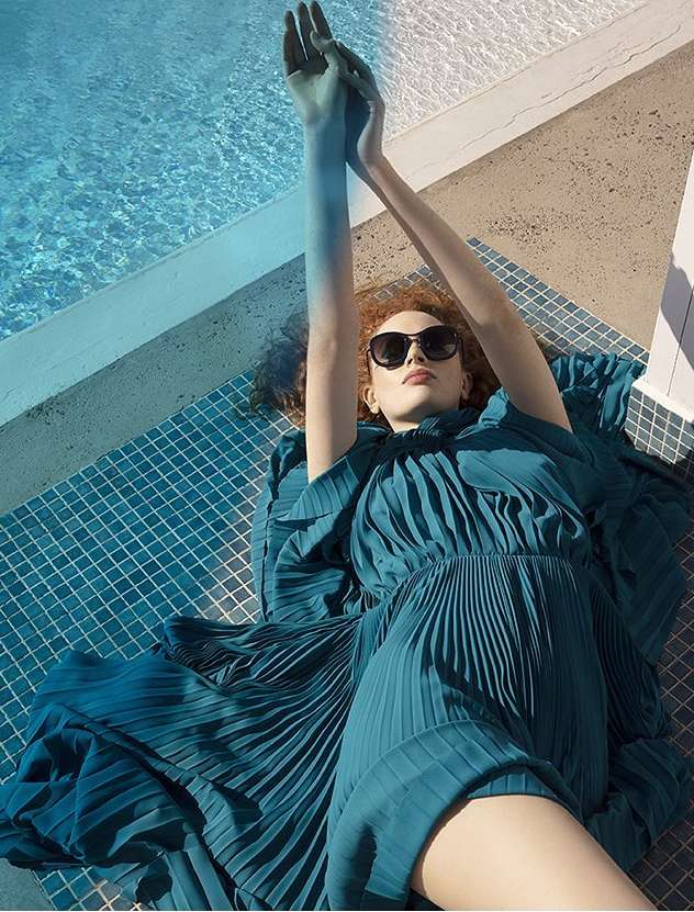 Woman with auburn hair in a blue dress and sunglasses reclines on blue mosaic tiles next to a pool with arms raised on a sunny day.