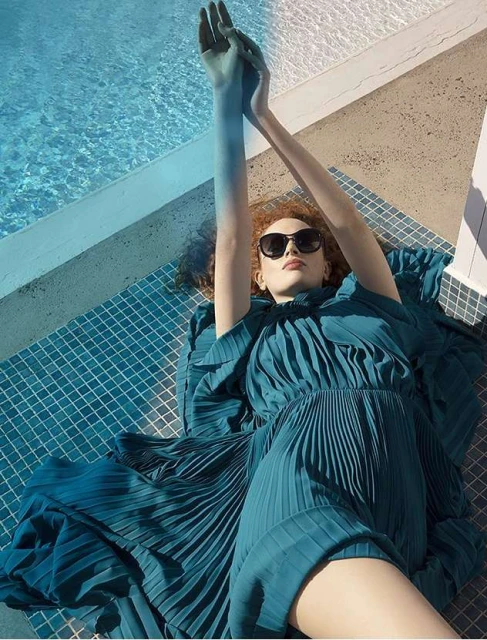 Woman with auburn hair in a blue dress and sunglasses reclines on blue mosaic tiles next to a pool with arms raised on a sunny day.