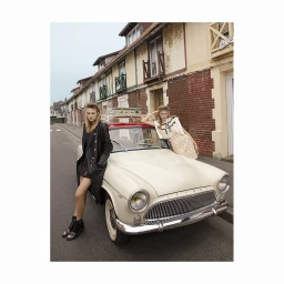 Two women posing with a vintage cream and red car on a narrow street lined with traditional buildings.
