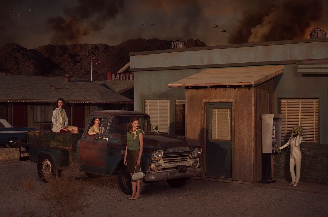 Three women and a figure in a white suit stand near a vintage truck and motel building against a desert and sky background.
