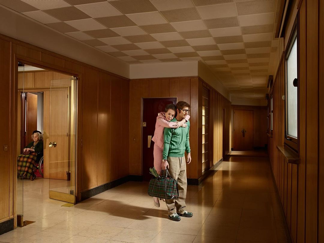 Young woman piggybacking a man in a hallway, with an elderly woman in a wheelchair visible through a doorway. Interior shot with warm lighting.