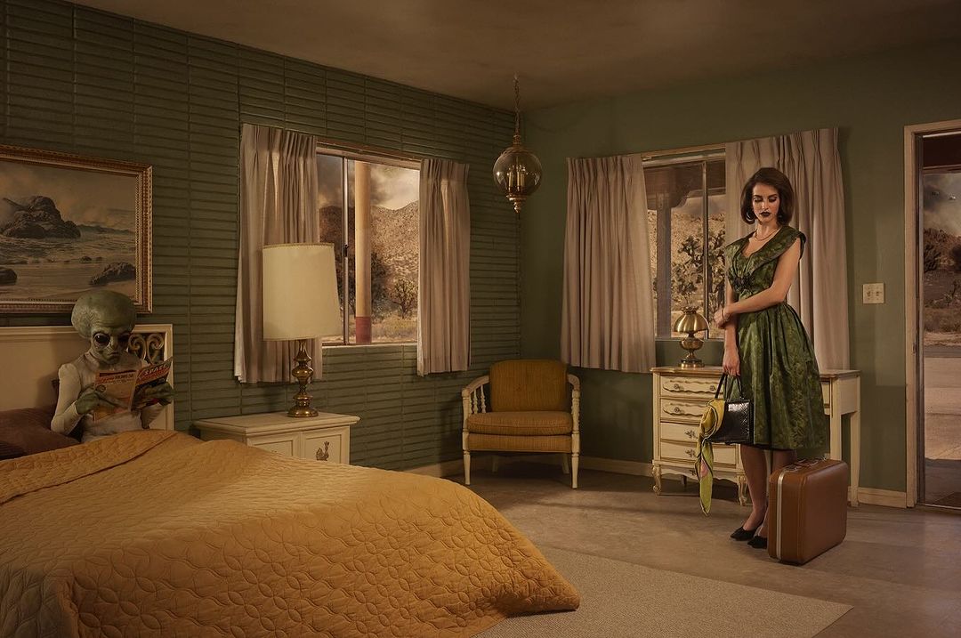 An alien sits in bed reading while a woman in a green dress stands near a suitcase, creating a retro science fiction scene in a motel.