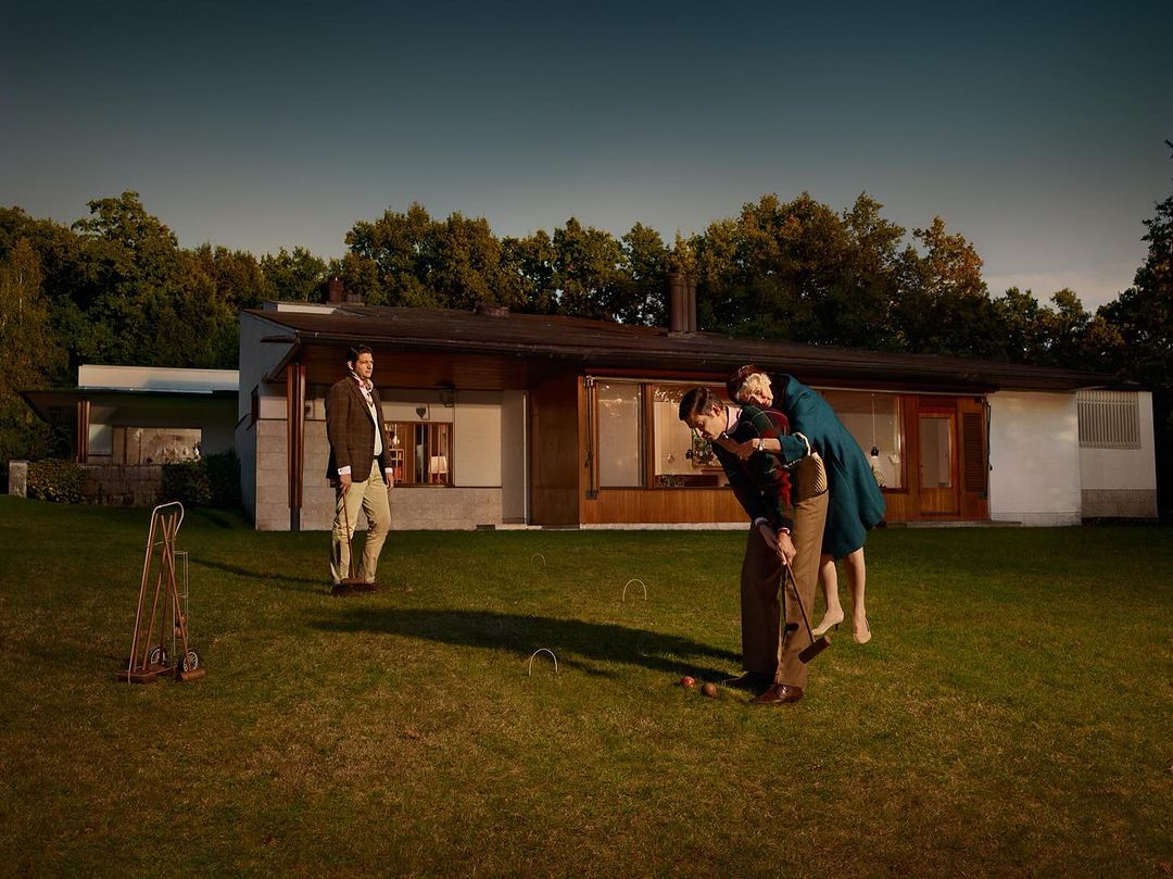 Man giving a piggyback ride to woman playing croquet on a lawn with another man standing nearby a modern house in daytime.