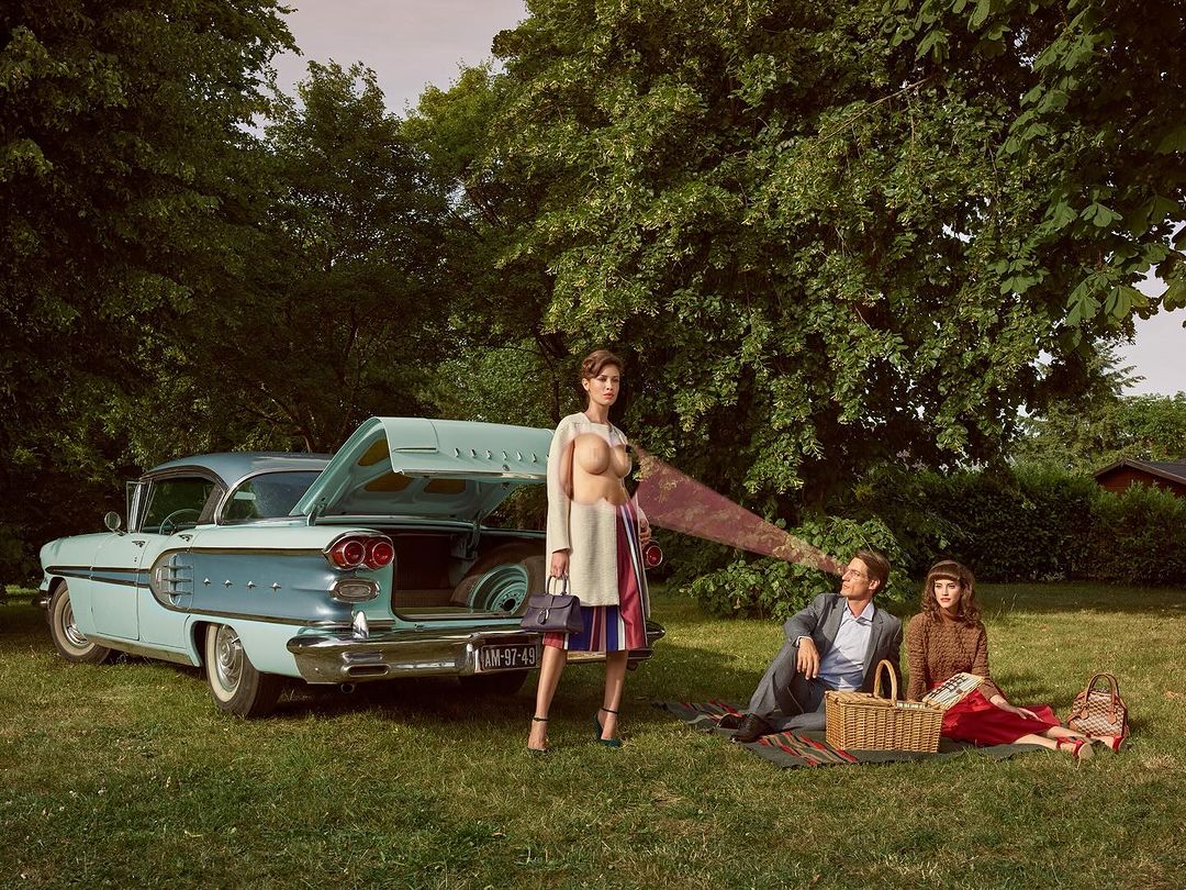 Vintage car, picnic scene with three people, including a partially nude woman. Outdoor setting with grassy ground and leafy trees.