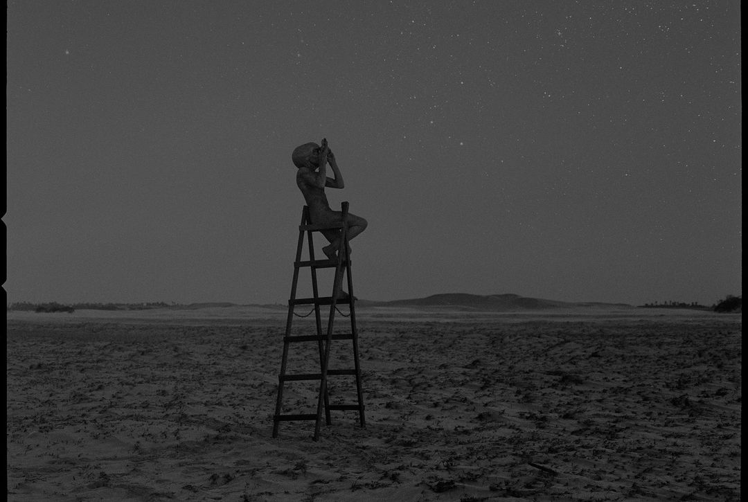 Monochromatic image depicts an individual seated on a ladder, looking upwards towards the starry night sky in an outdoor setting.