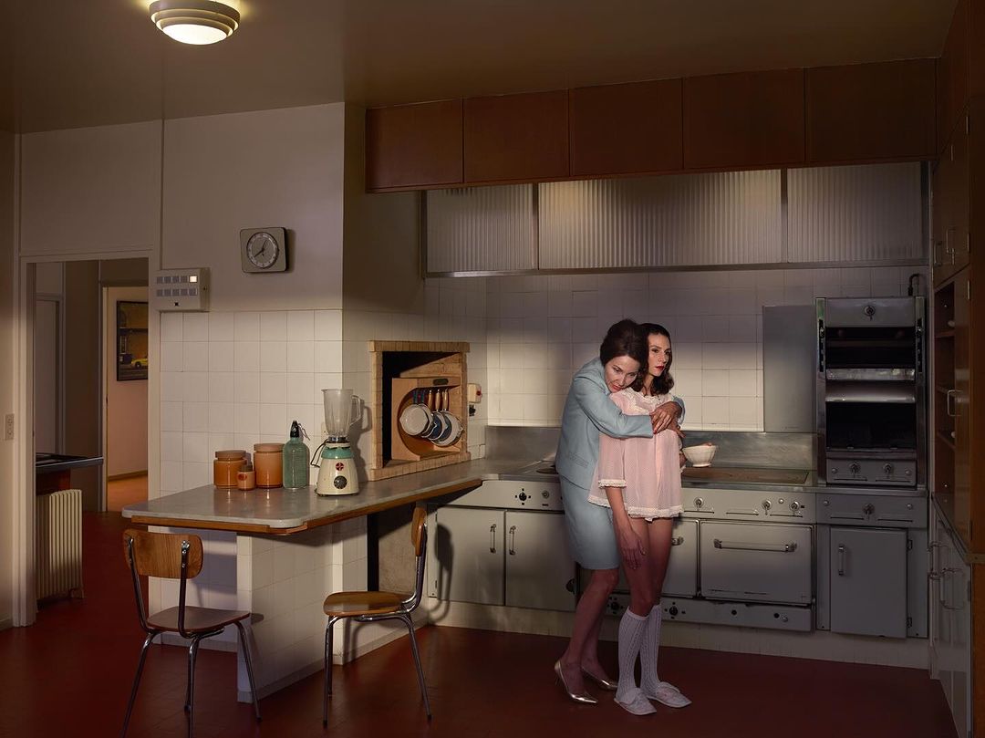 Two women embrace in a retro-style kitchen. One woman in a light blue suit hugs a younger woman wearing a pink nightgown and knee socks.