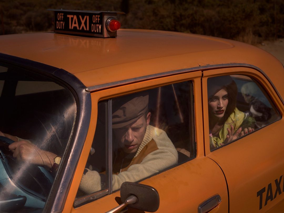 A vintage yellow taxi cab with a man driving and woman passenger looking out the window in a retro-styled outdoor scene.