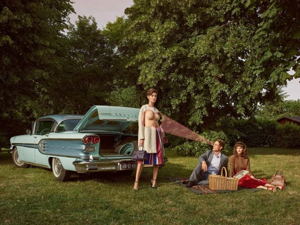 Woman standing next to a vintage light blue car with open trunk near a man and woman seated on picnic blanket on grass