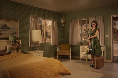 A woman in a green dress standing with luggage next to an alien reading a magazine in bed inside a retro bedroom.