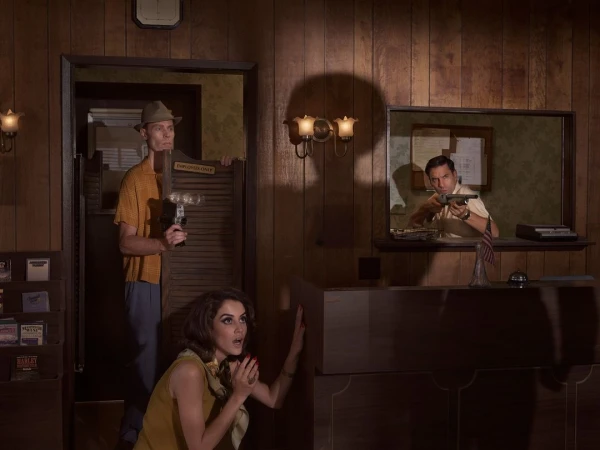 Three people in a retro motel lobby with wood paneled walls, featuring a man holding a gun behind a counter, a woman crouching anxiously, and another man holding a camera near a doorway.