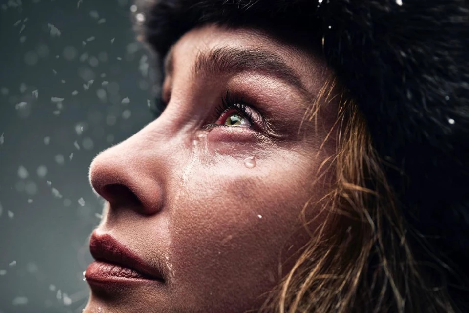 Close-up of a woman with a tear on her cheek and water droplets on her face, wearing a dark fur hat and looking up.