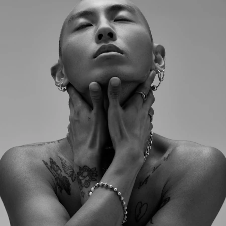 Black-and-white close-up studio shot of an Asian adult male with a shaved head, hands on neck, earrings, and various tattoos.