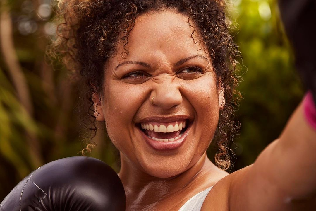 Close up of a smiling woman with curly hair wearing boxing gloves and workout attire against a leafy green background, captured in natural light.