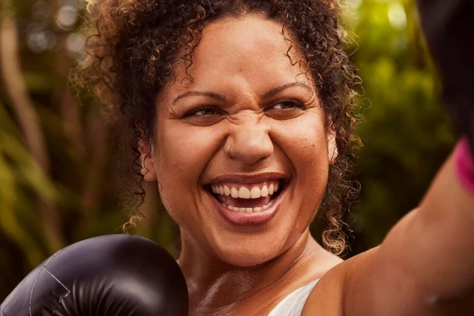 Close up of a smiling woman with curly hair wearing boxing gloves and workout attire against a leafy green background, captured in natural light.